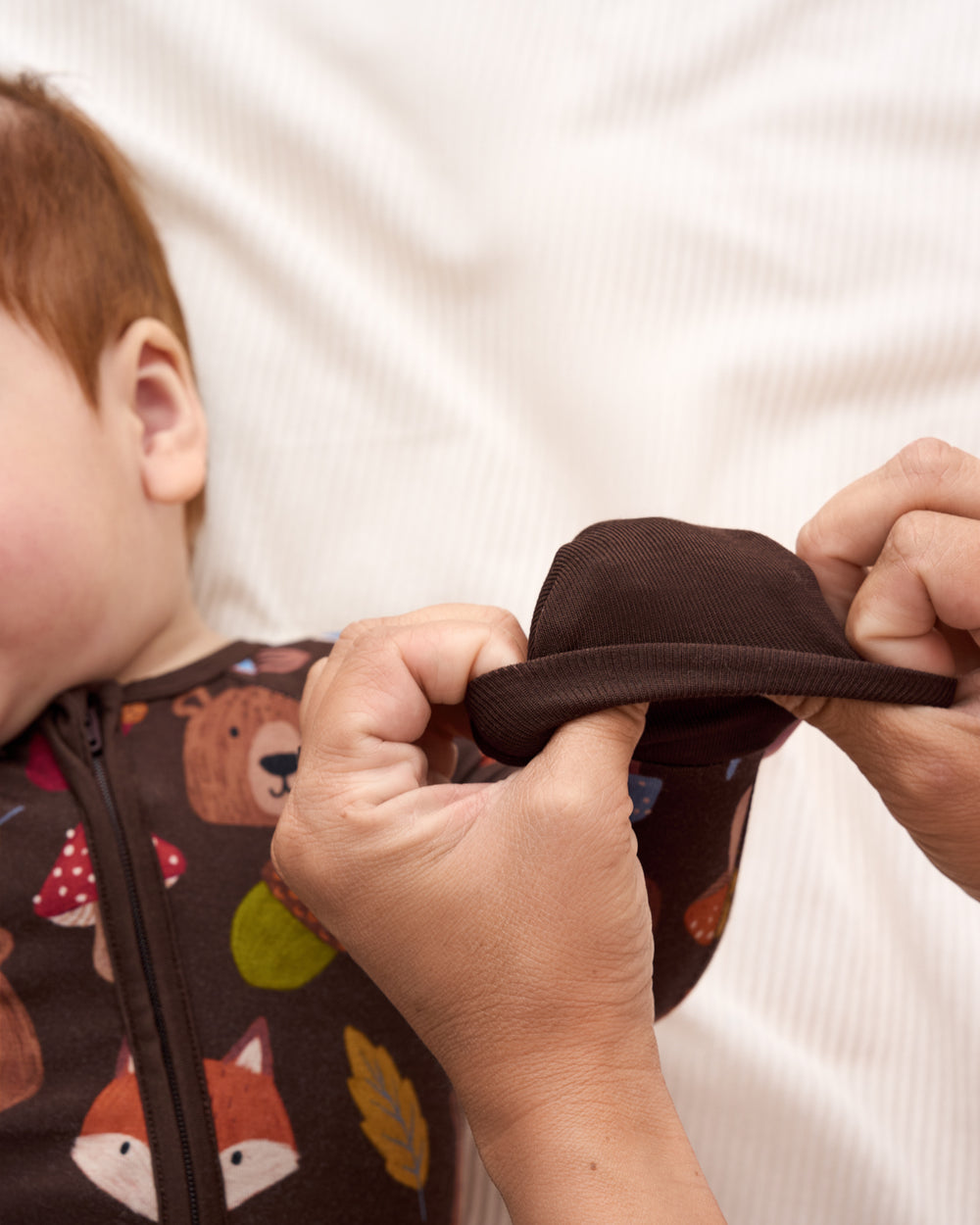 A close-up lifestyle shot of a baby wearing a brown zip-up pajama with a woodland animal print, while an adult's hands adjust the sleeve.