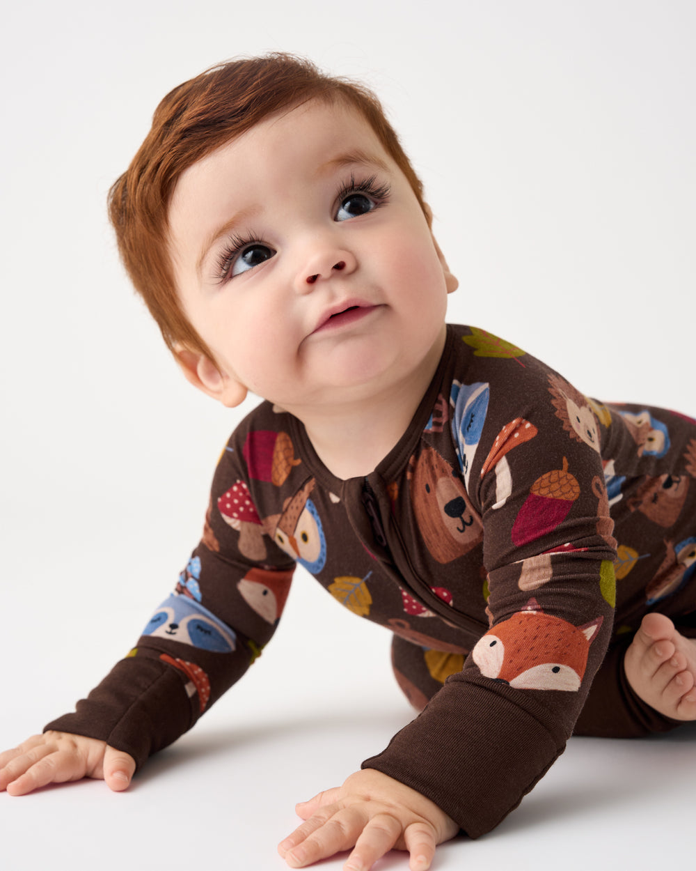 A baby crawling on a white studio floor wearing a long-sleeve brown pajama top with a colorful woodland animal print including foxes, bears, and acorns.