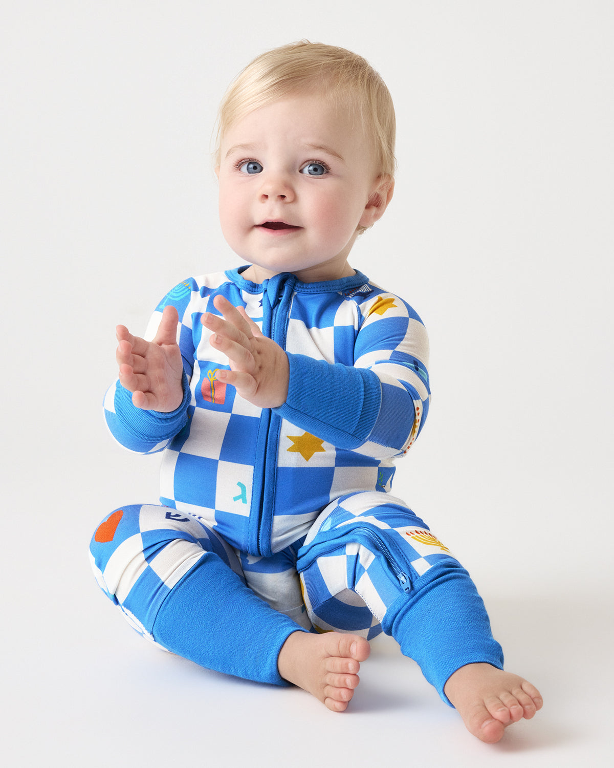 A baby sitting on a light gray studio background wearing a blue and white checkered zip-up one-piece pajama with colorful icons.