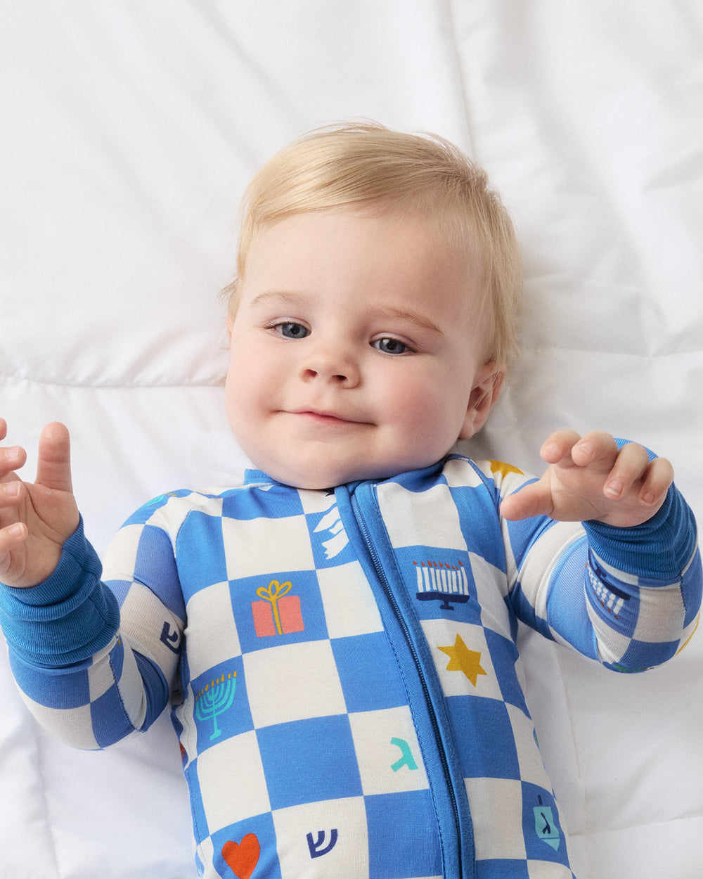 A baby lying on a white quilted blanket, wearing a blue and white checkered zip-up sleeper decorated with small colorful symbols.
