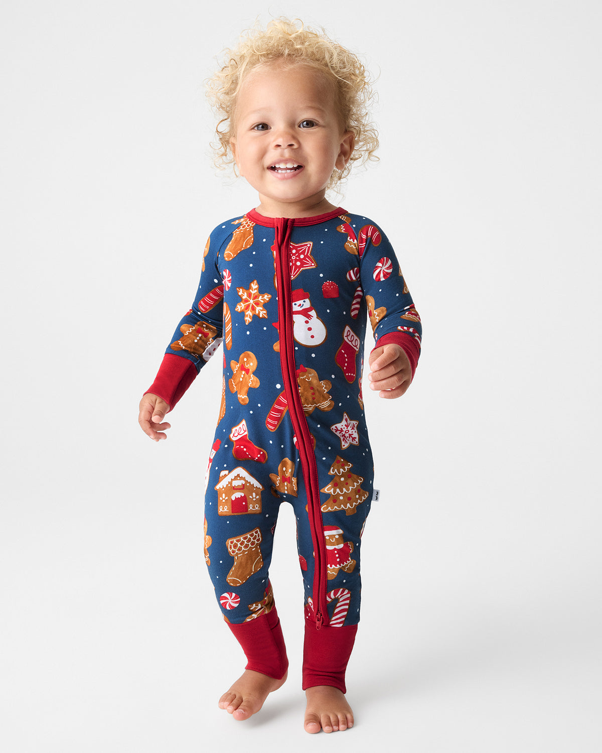 A toddler stands barefoot on a light gray studio background, wearing a navy blue one-piece pajama with a red zipper and cuffs, and a holiday print featuring gingerbread cookies, Santa Claus, candy canes, snowman, and stars.