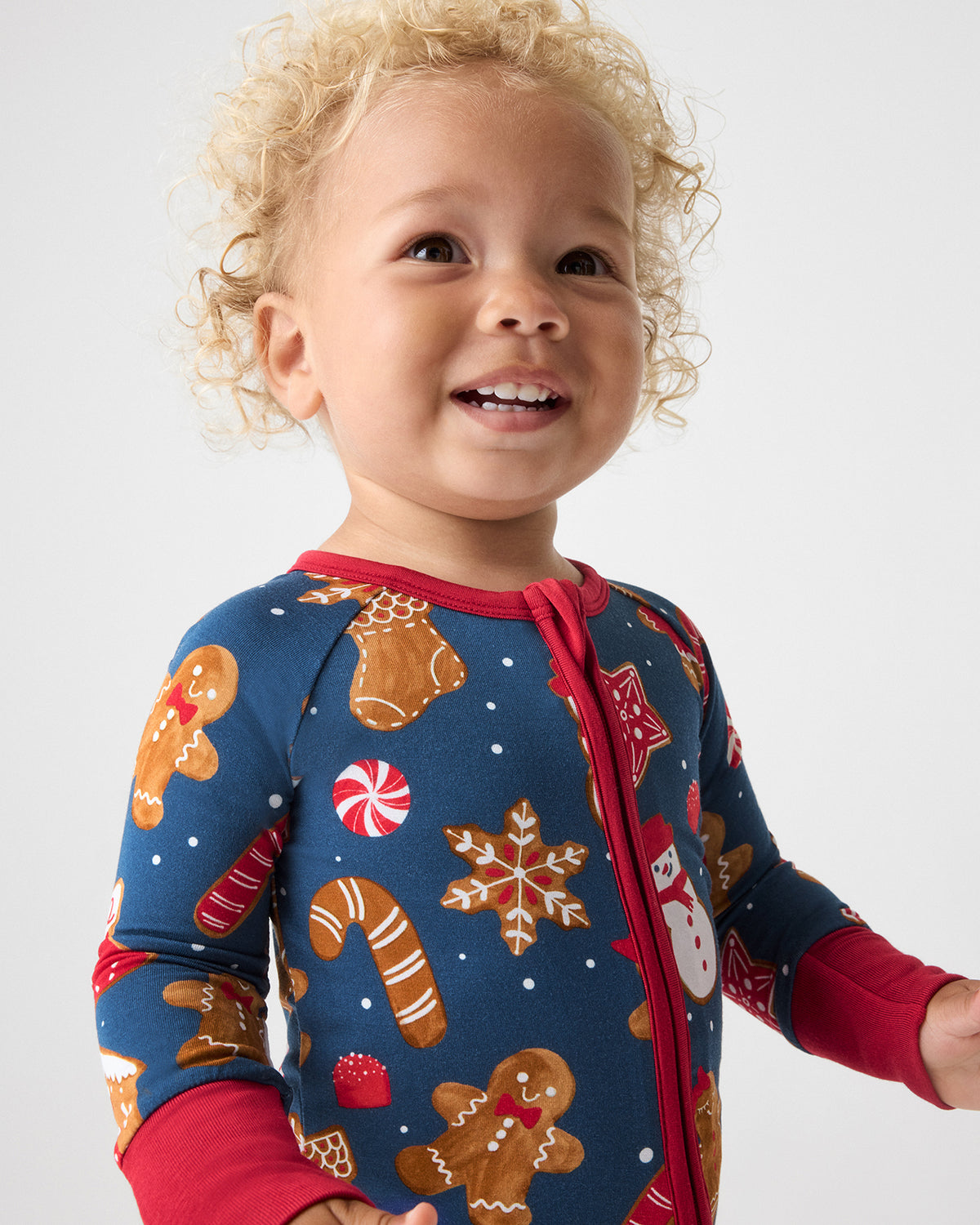 A smiling toddler wearing a festive blue long-sleeve pajama top with red trim and gingerbread-themed prints, photographed against a light gray studio background.