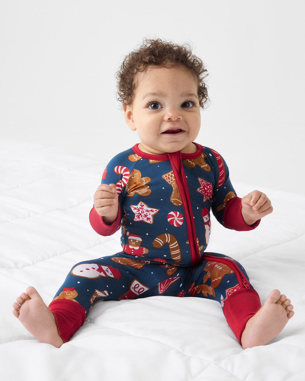 A baby sitting on a white quilted bed wearing a navy blue zip-up pajama with a festive holiday print of gingerbread cookies, candy canes, snowmen, and stars, with red cuffs and collar.
