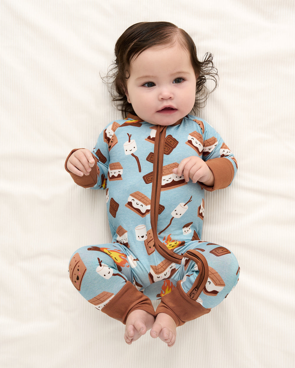 A baby lying on a white textured blanket, wearing a light blue footless zip-up sleeper with a brown and white s'mores print.
