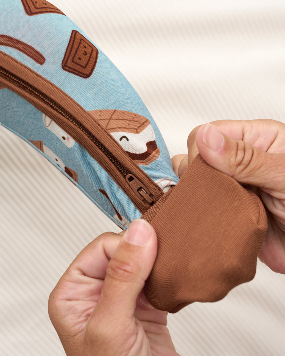 Close-up of a person's hands demonstrating the stretchy, elastic brown fabric of a zipper pouch. The pouch features a light blue background with a whimsical pattern of brown chocolate squares and white marshmallows.
