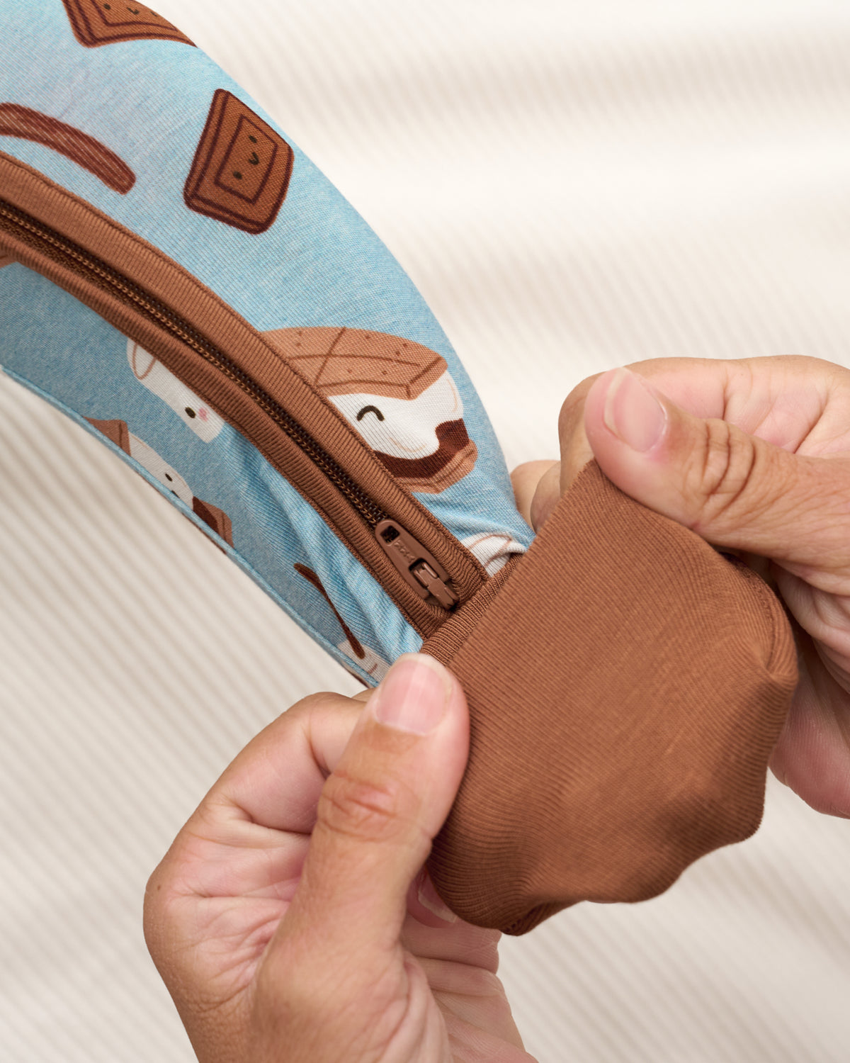 Close-up of a person's hands demonstrating the stretchy, elastic brown fabric of a zipper pouch. The pouch features a light blue background with a whimsical pattern of brown chocolate squares and white marshmallows.