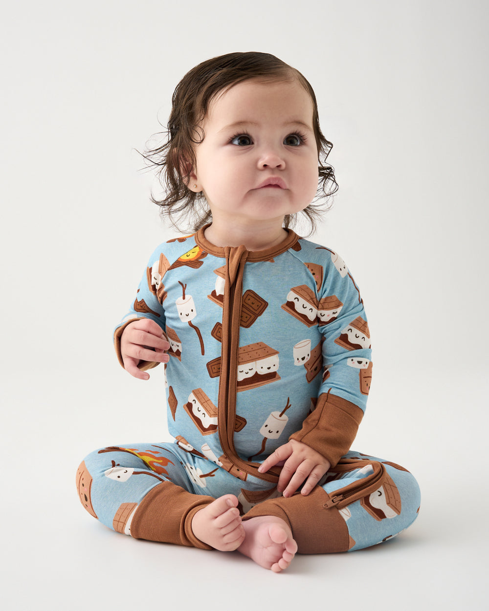 A baby sitting cross-legged wearing a light blue one-piece pajama with a brown zipper and brown cuffs, legs, and collar, decorated with a repeating pattern of gummy bears and marshmallows, on a light gray studio background.