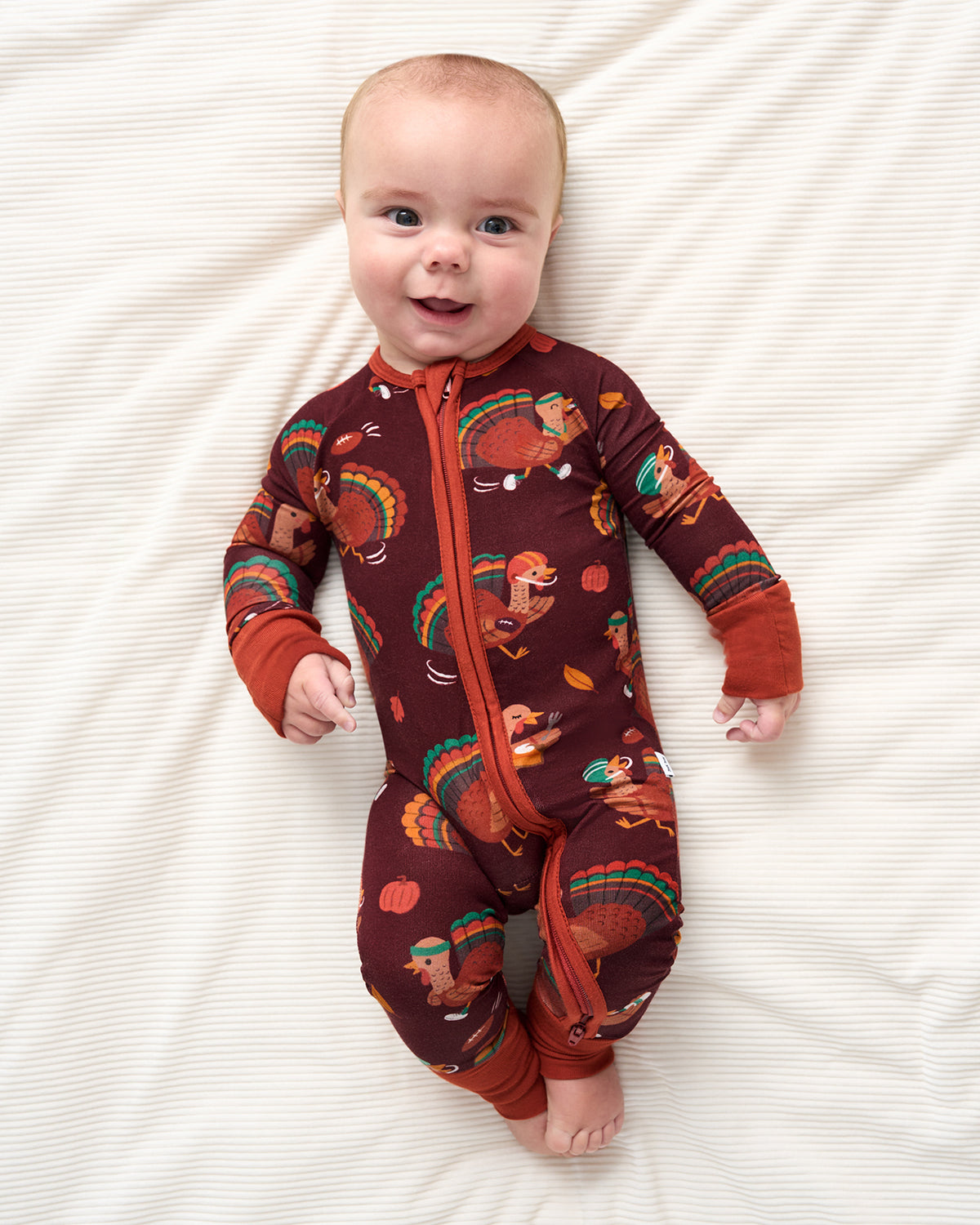 A baby lying on a cream-colored textured blanket, wearing a long-sleeve, one-piece zip-up sleeper in a dark brown color with a colorful autumn turkey and pumpkin pattern.