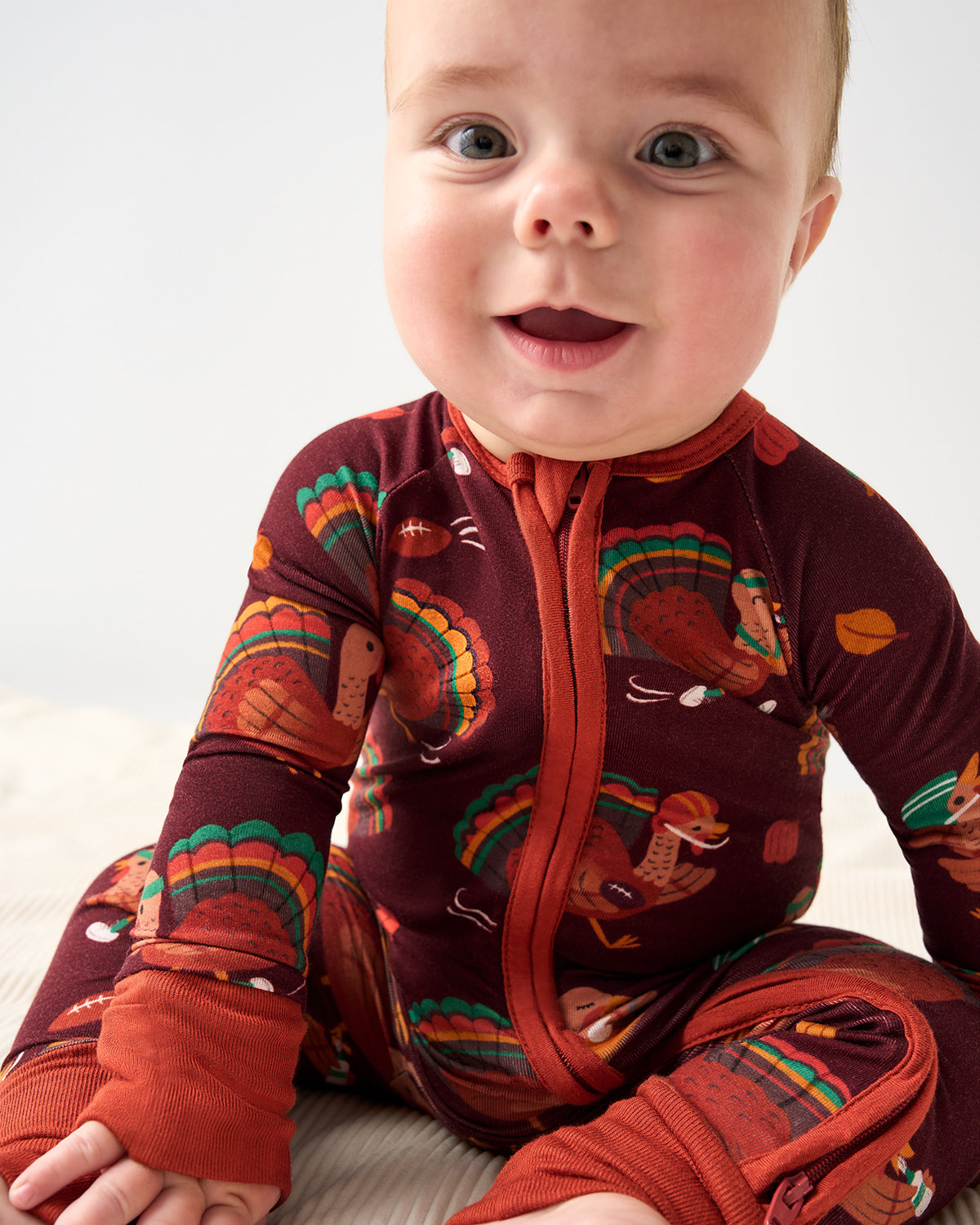 A baby wearing a long-sleeve, one-piece zip-up sleeper with a Thanksgiving turkey print, sitting against a plain light background.