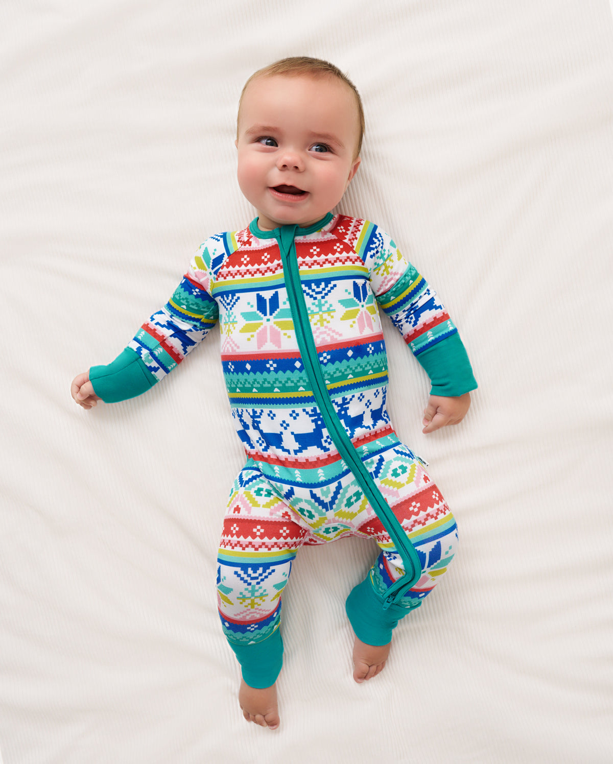 A baby lying on a white textured blanket wearing a colorful long-sleeve zip-up sleeper with a festive Fair Isle pattern in red, blue, green, yellow, and white.