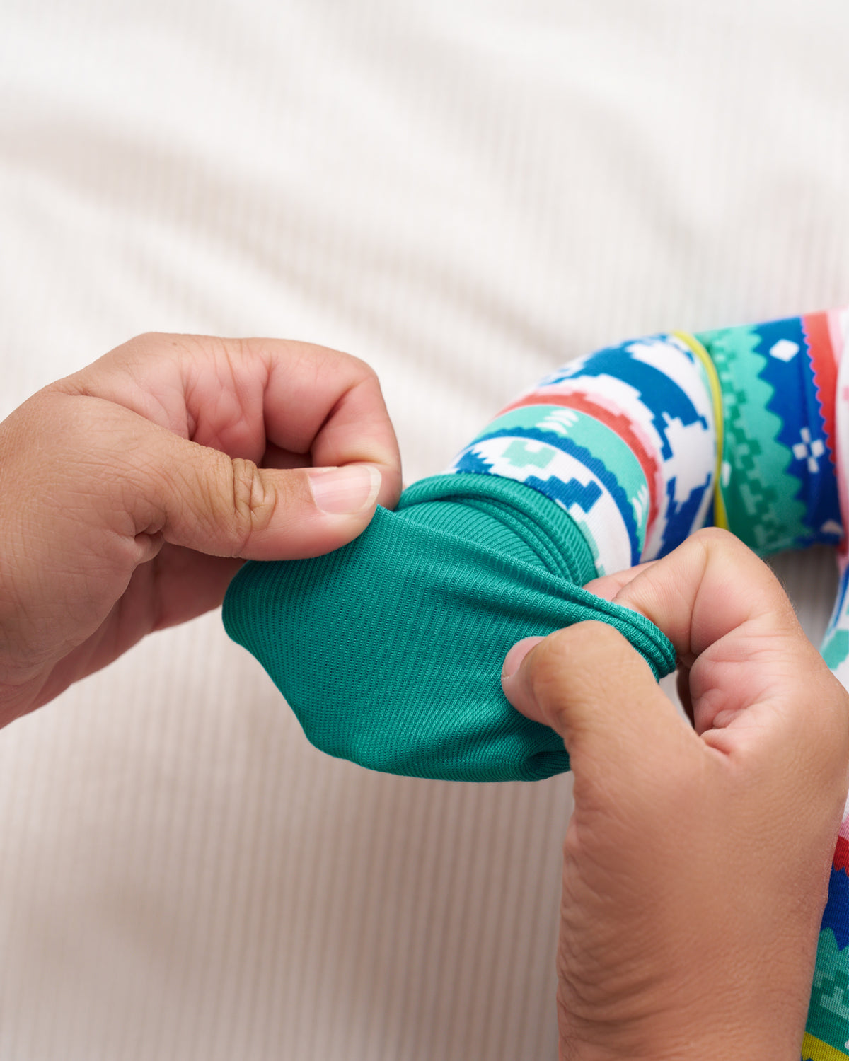 Close-up of a person's hands stretching a teal green fabric sleeve on a colorful patterned PJ against a white ribbed fabric background.