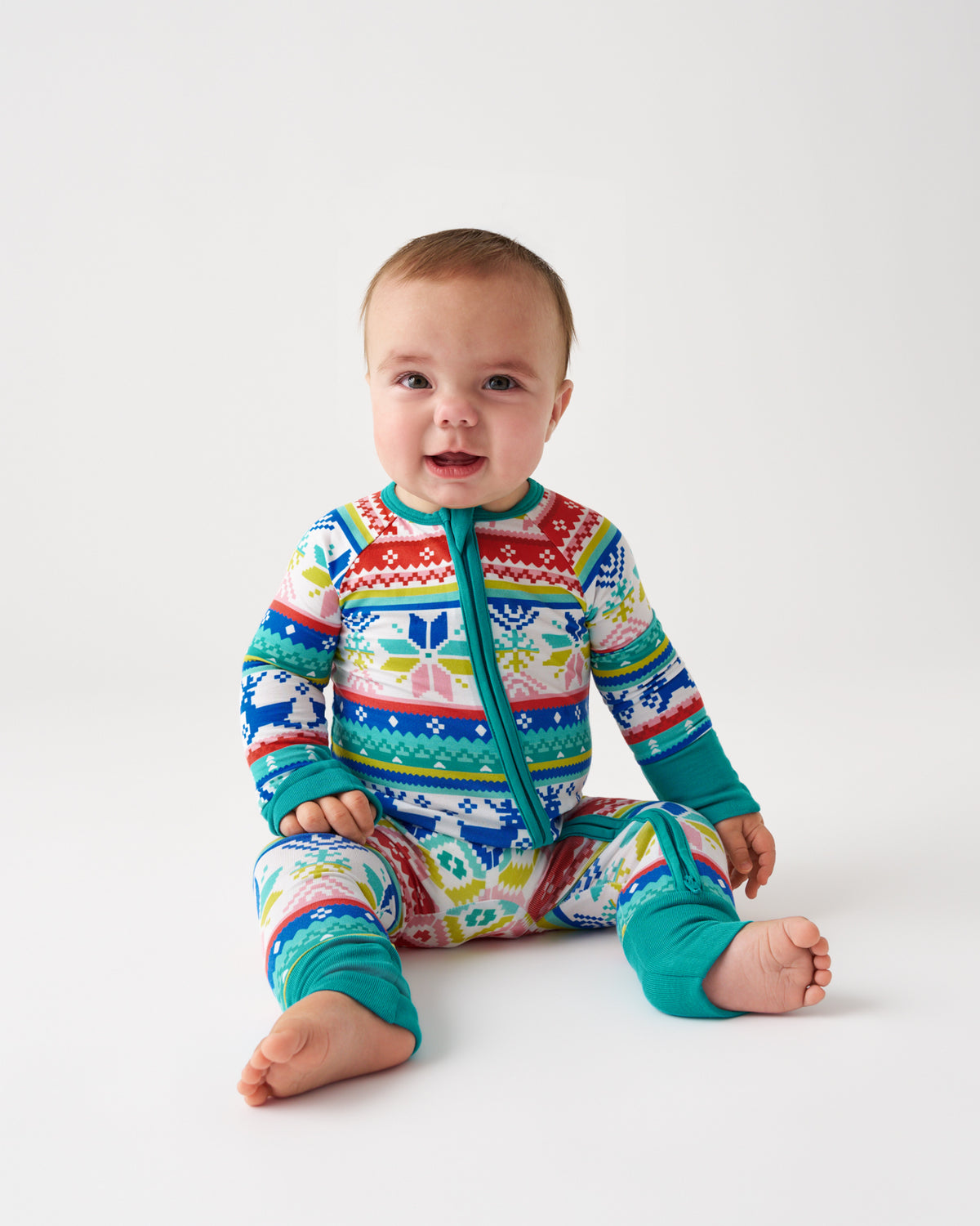 A baby sitting on a light gray studio background wearing a colorful long-sleeve zip-front footed sleeper with a multicolor Fair Isle pattern and teal cuffs.