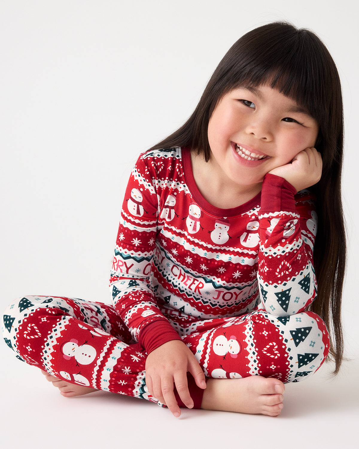 A young girl with long dark hair sits cross-legged on a light gray studio background, wearing a red and white holiday-themed long-sleeve pajama set with snowman and Christmas patterns.