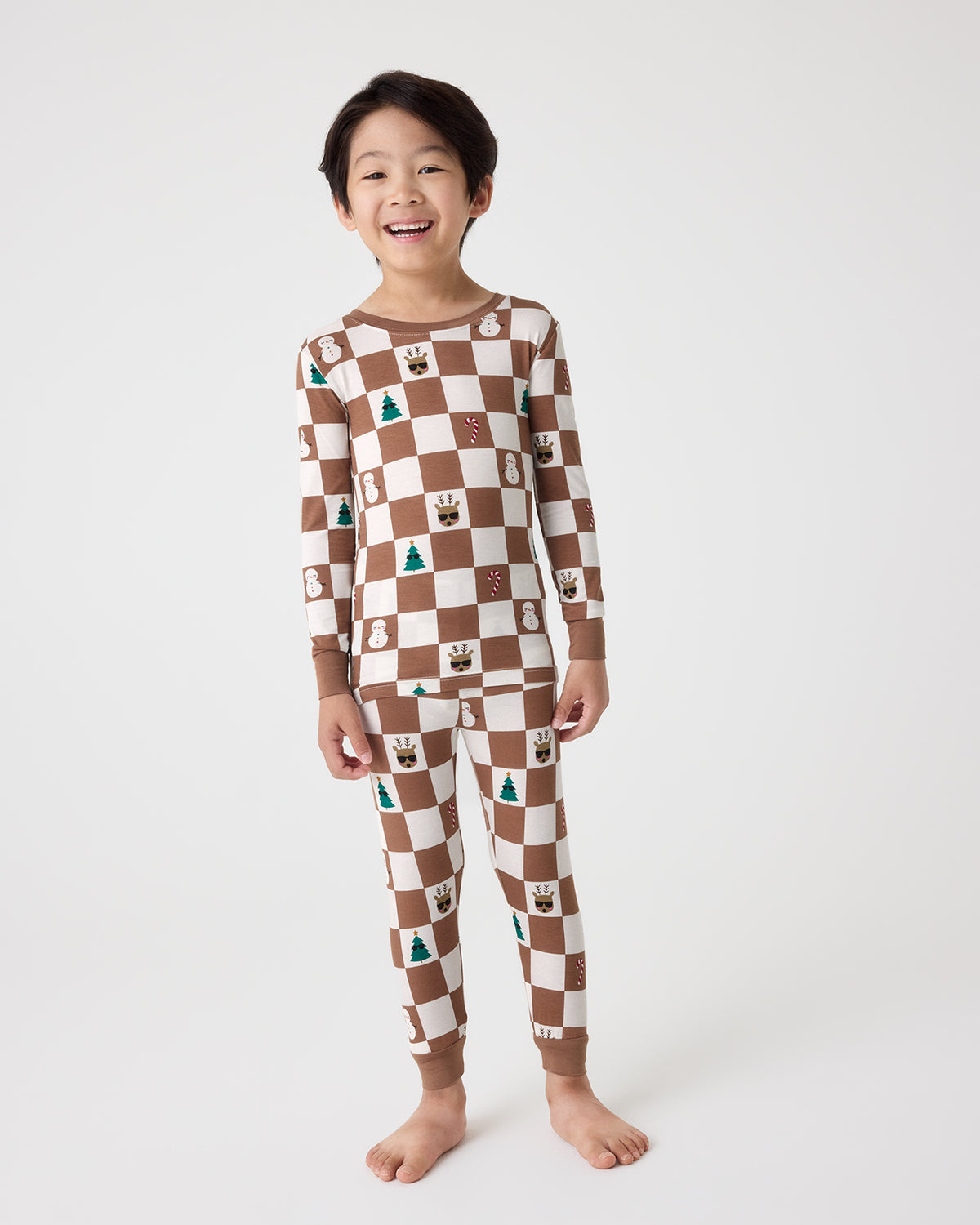 A young child wearing a brown and cream checkered long-sleeve pajama set with small holiday-themed illustrations, standing against a plain light gray studio background.