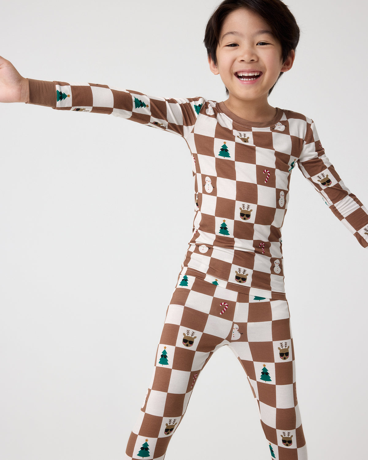 A smiling young boy wearing a brown and cream checkerboard long-sleeve pajama set with small holiday motifs, posed against a light gray studio background.