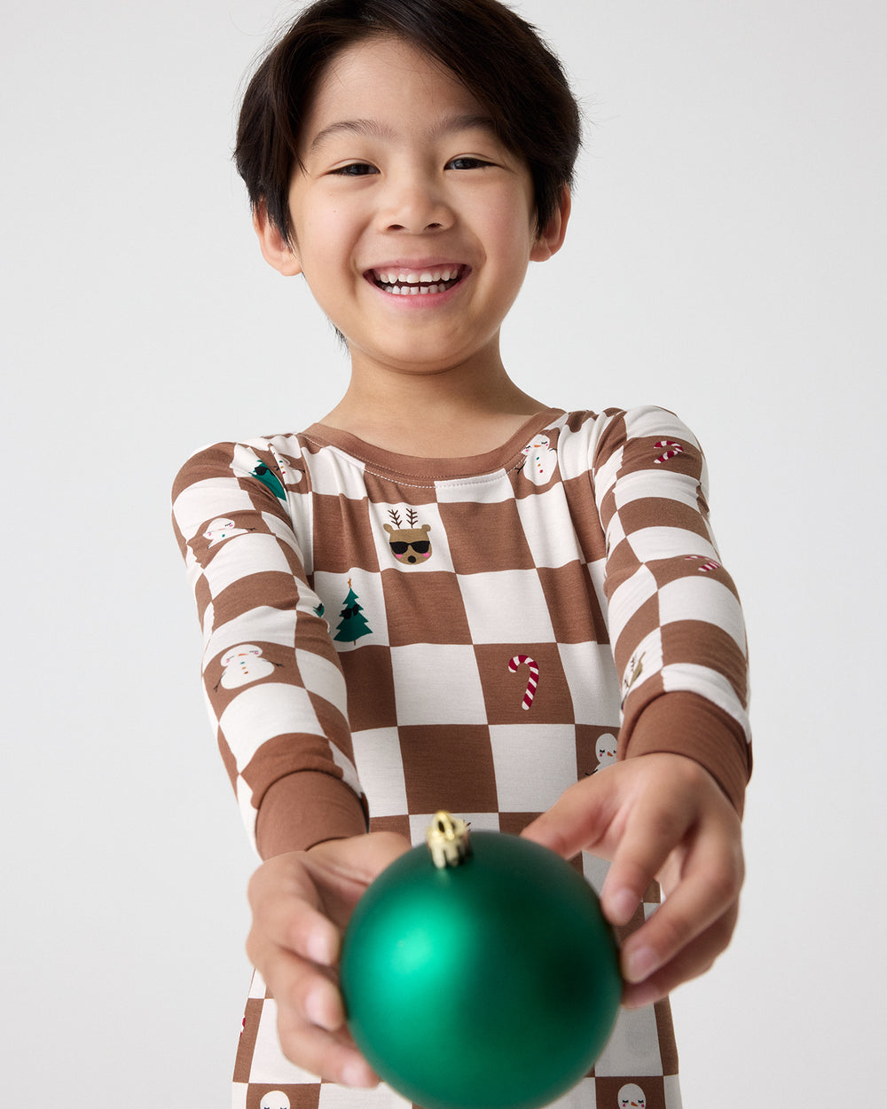 A smiling young boy wearing a brown and cream checkered long-sleeve pajama shirt with small holiday motifs, holding a shiny green Christmas ornament against a light gray background.