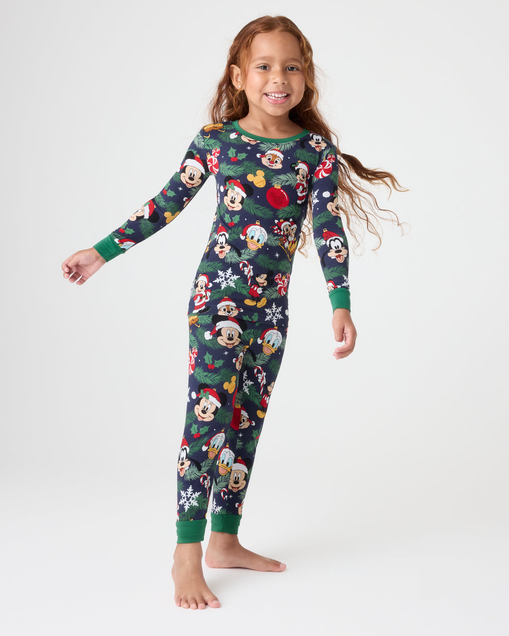 A young girl stands barefoot against a light gray studio background, wearing a long-sleeve full-body pajama set with a dark green base and an all-over festive character print including Mickey Mouse, Donald Duck, and other holiday motifs.
