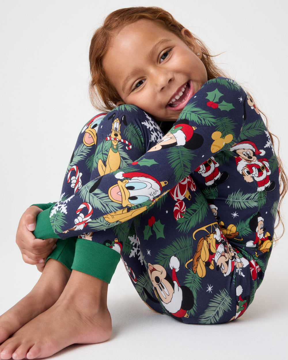 A young girl smiling and crouching while wearing a navy blue Christmas-themed pajama set with a colorful Disney character print and green cuffs, photographed against a light gray studio background.