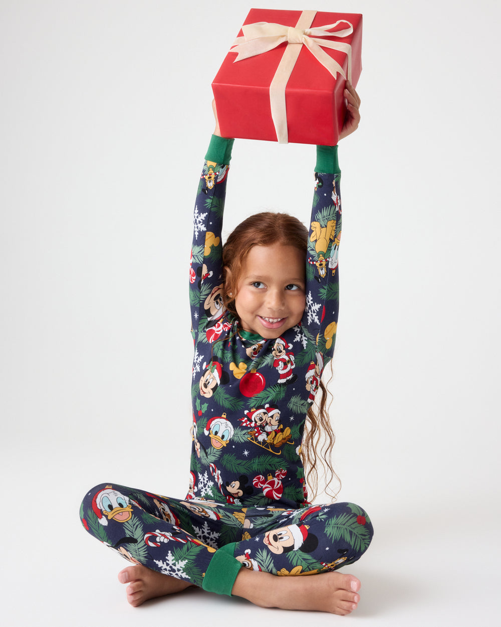 A young girl sits cross-legged on a white studio background, holding a red gift box above her head. She is wearing dark green Christmas-themed pajamas with a colorful cartoon character print.
