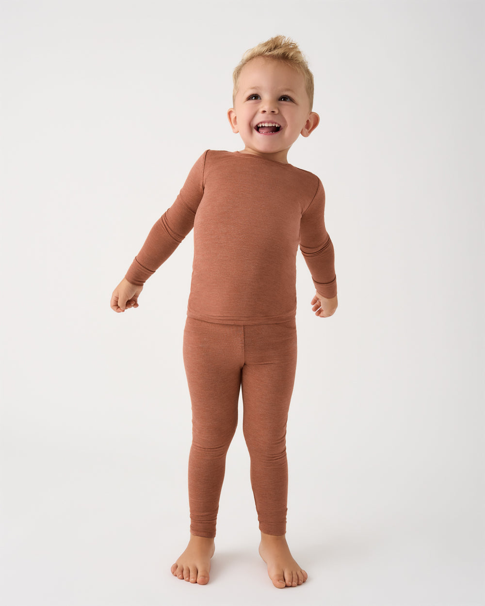 A smiling toddler standing barefoot against a plain white studio background, wearing a matching long-sleeve top and leggings set in a warm rust or terracotta color.