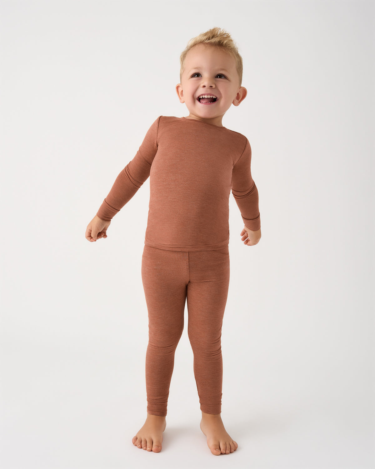 A smiling toddler standing barefoot against a plain white studio background, wearing a matching long-sleeve top and leggings set in a warm rust or terracotta color.