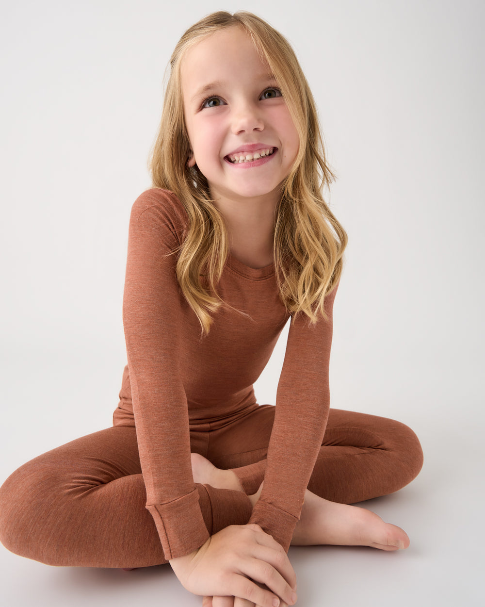 A young girl with blonde hair sits cross-legged on a light gray studio background, wearing a matching rust-brown long-sleeve top and leggings set.