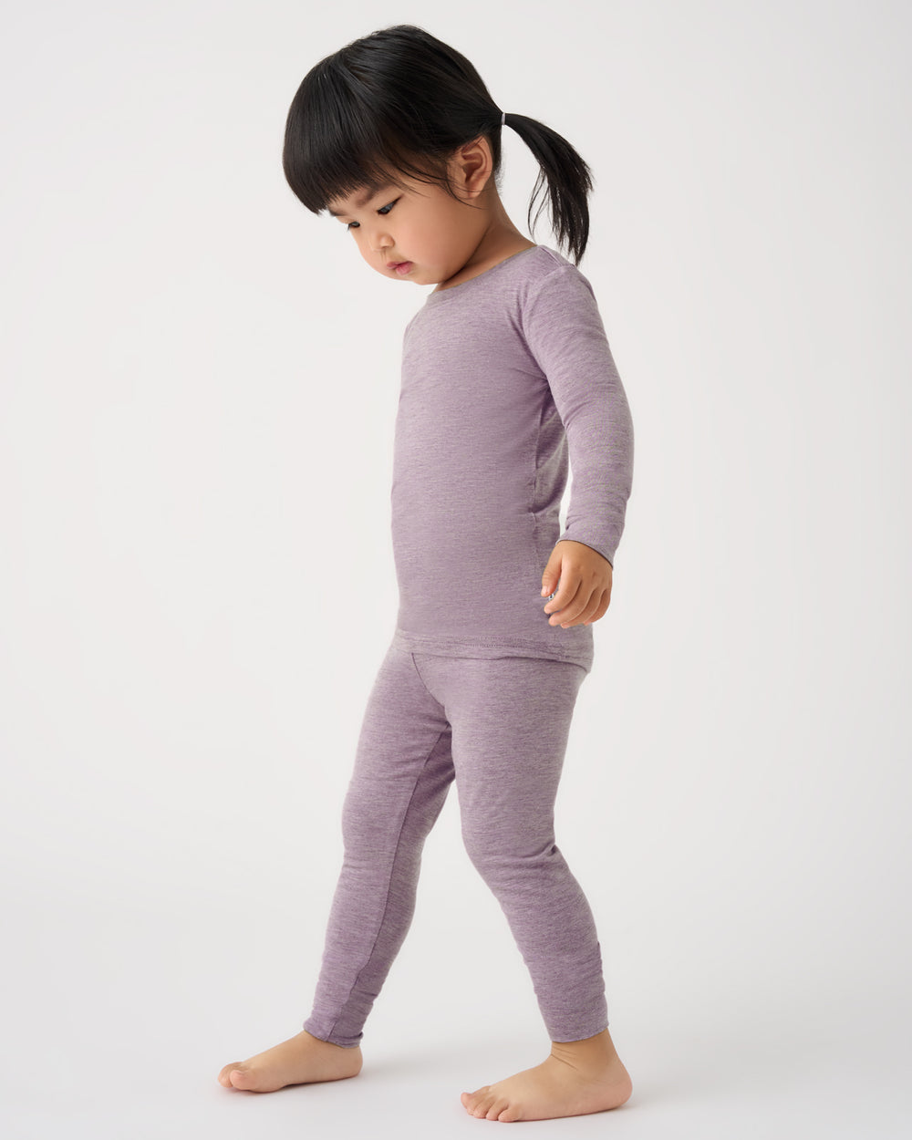 A toddler girl wearing a matching two-piece long-sleeve pajama set in a heathered light purple/lavender color, standing against a plain white studio background.