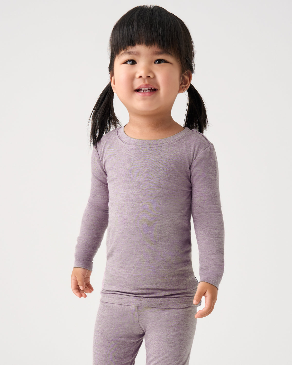 A young girl with dark hair in pigtails wearing a light purple long-sleeve crewneck shirt and matching pants, standing against a plain light gray background.