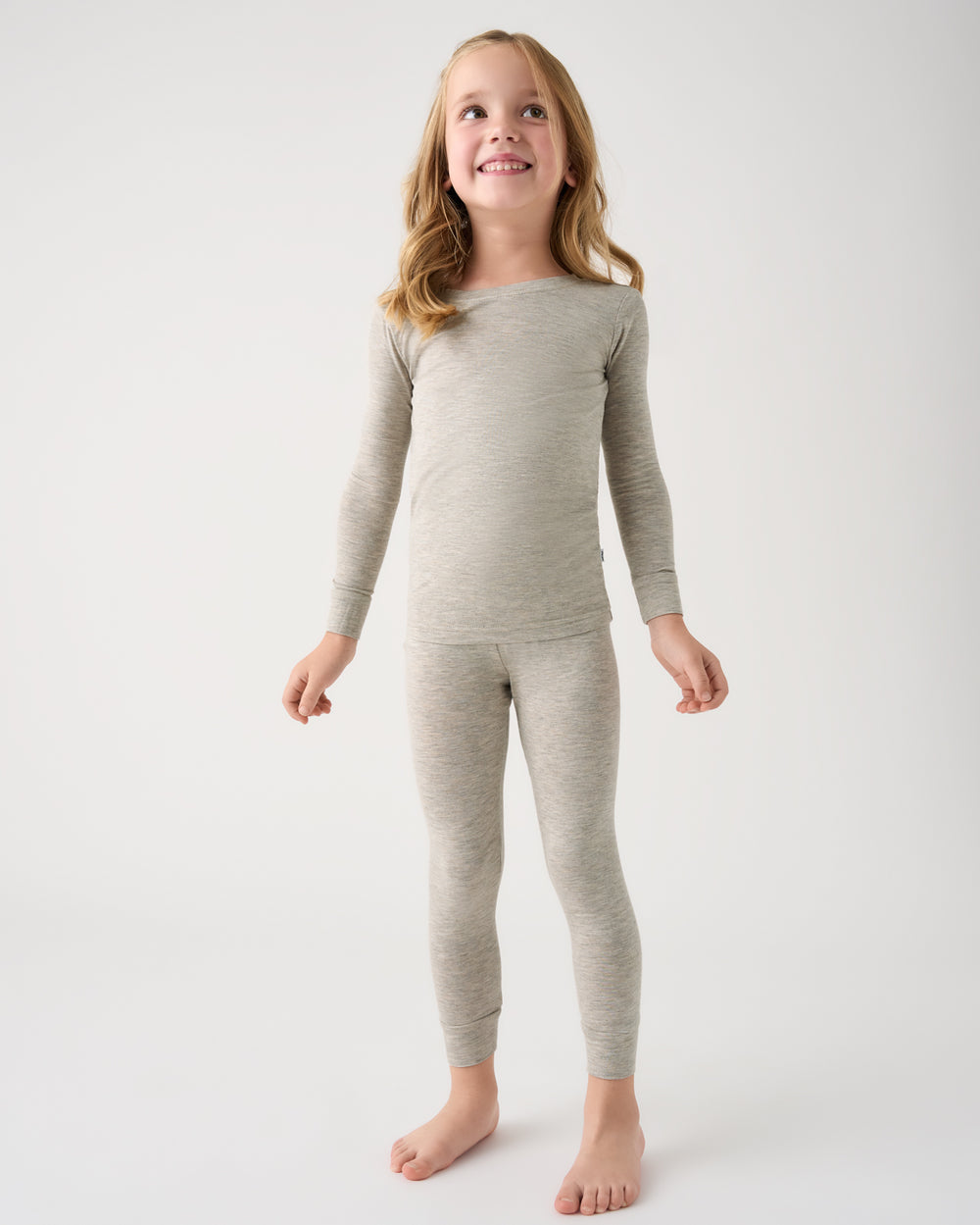 A young girl with blonde hair stands barefoot against a light gray studio background, wearing a matching light gray long-sleeve top and leggings set.