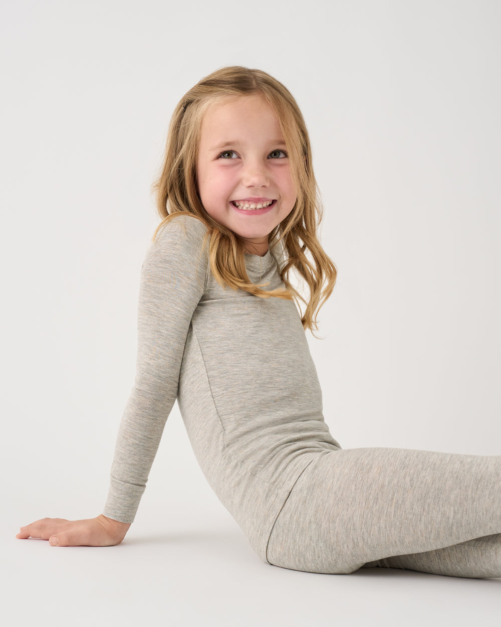 A young girl with blonde hair sits on a white studio floor wearing a light heather gray long-sleeve turtleneck jumpsuit.