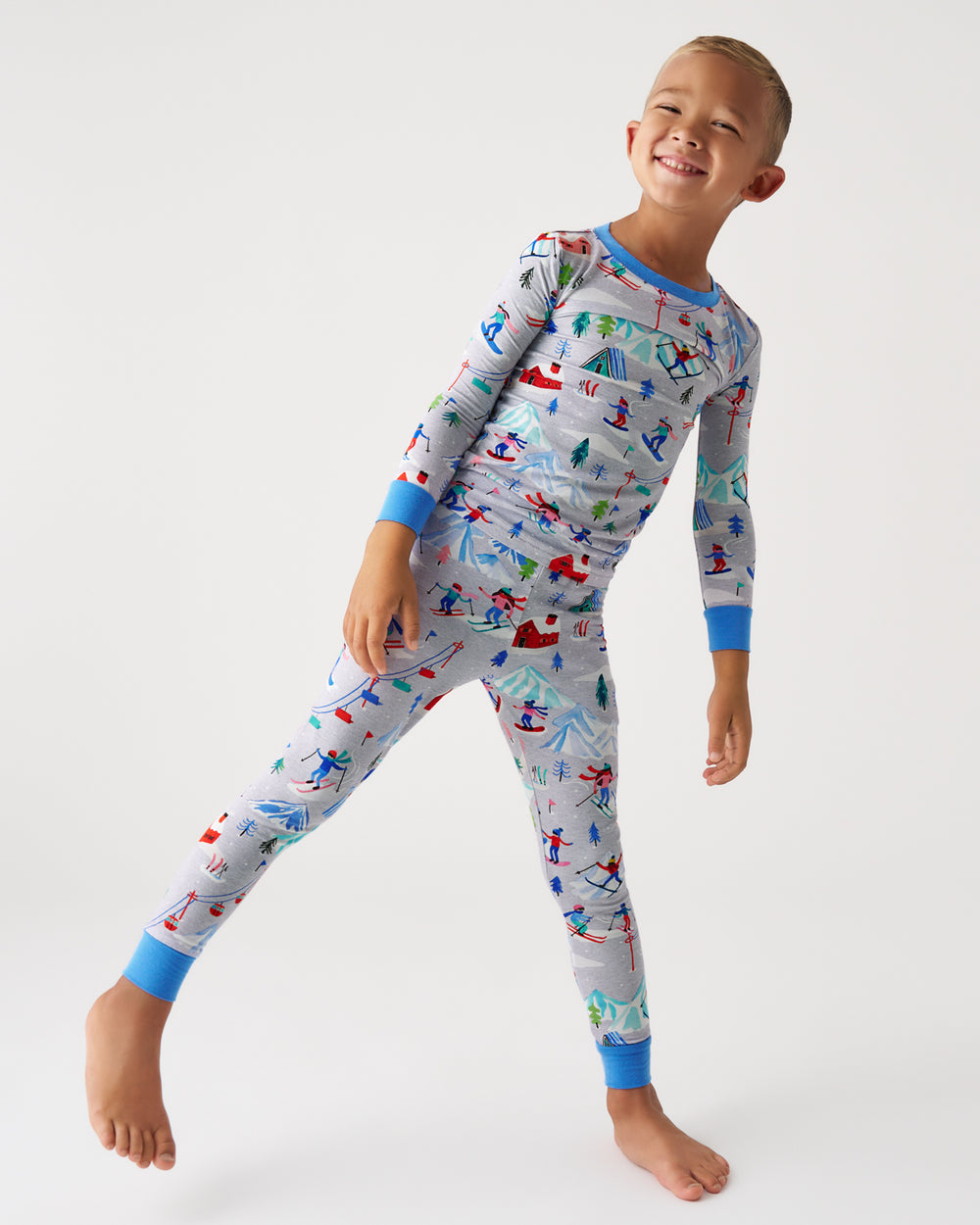 A smiling young boy wearing a long-sleeve pajama set with a colorful winter sports print on a light gray background, posed against a plain white studio background.