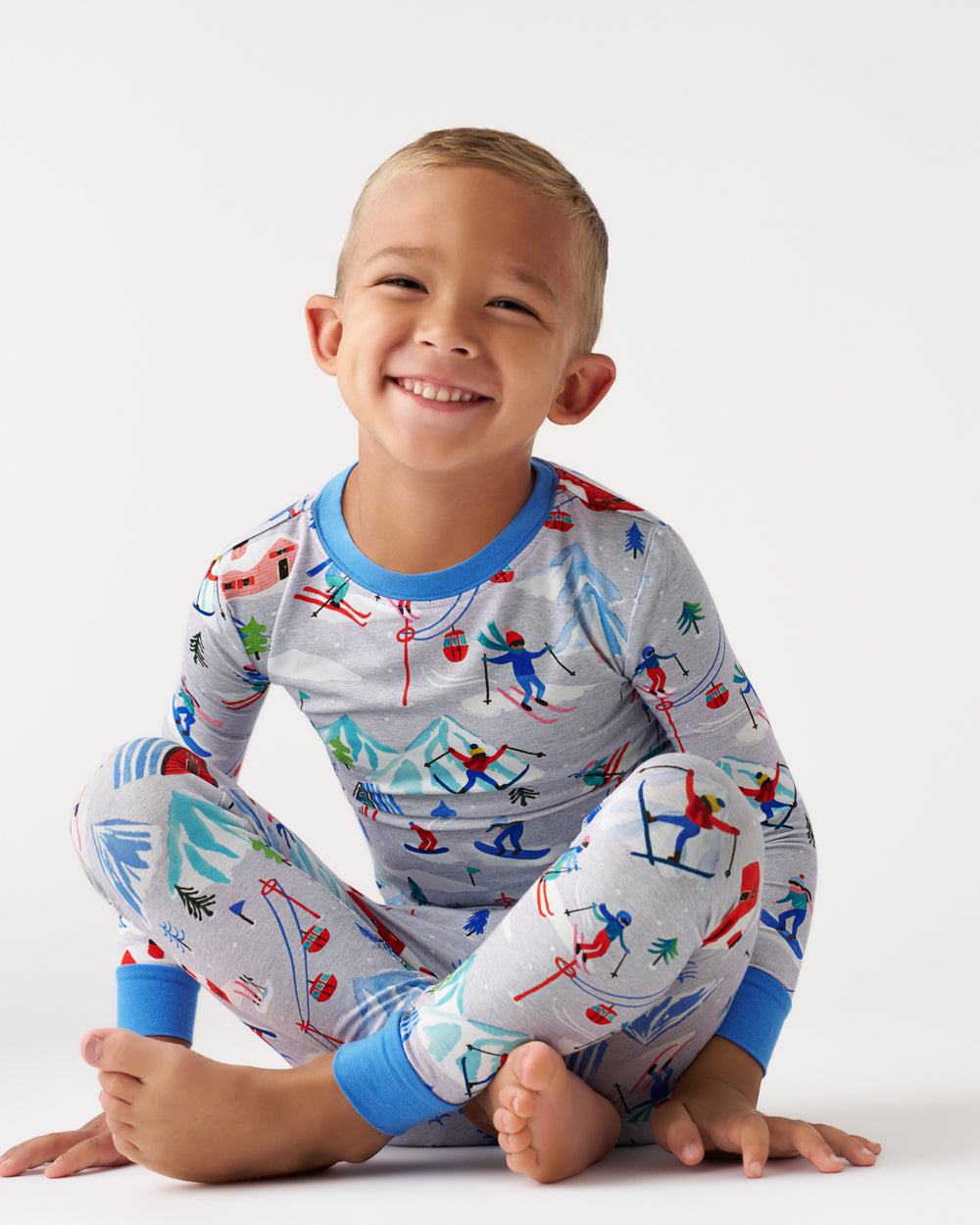 A smiling young boy wearing a long-sleeve pajama set with a colorful ski and snowboard pattern on a light gray background, sitting cross-legged against a plain white studio backdrop.