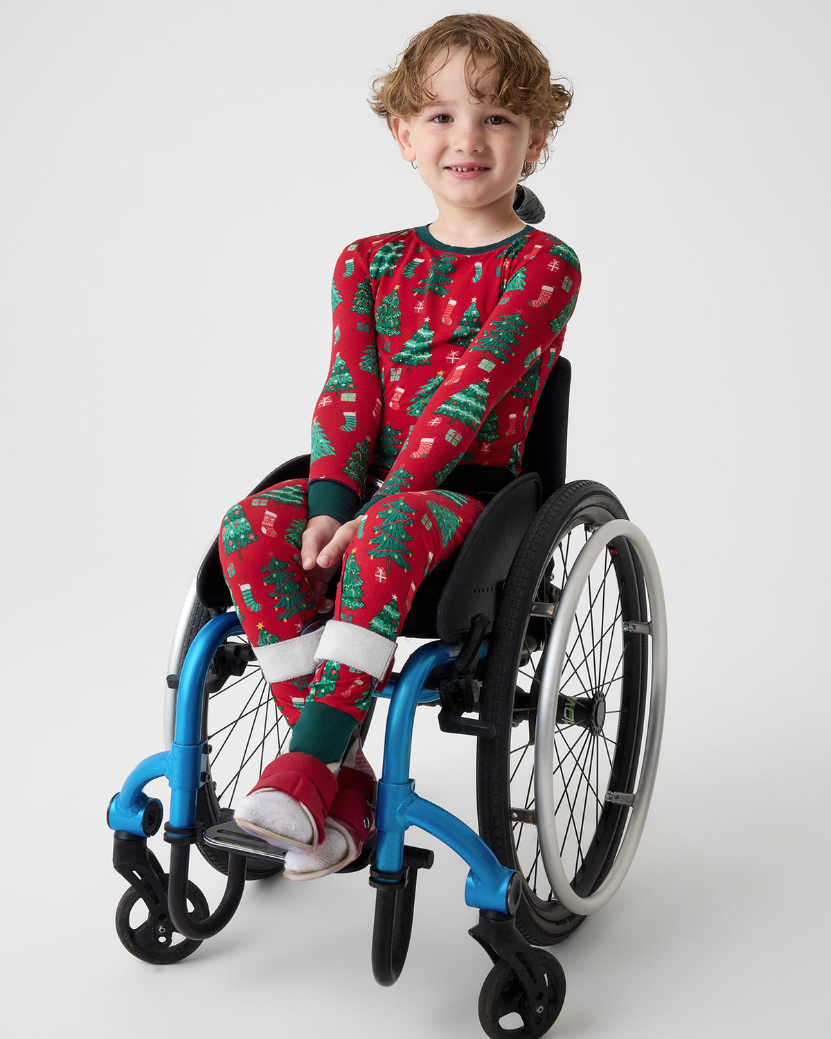A young child sitting in a wheelchair, wearing a festive red and green Christmas-themed long-sleeve pajama set with a tree pattern. The child is photographed against a light gray studio background.