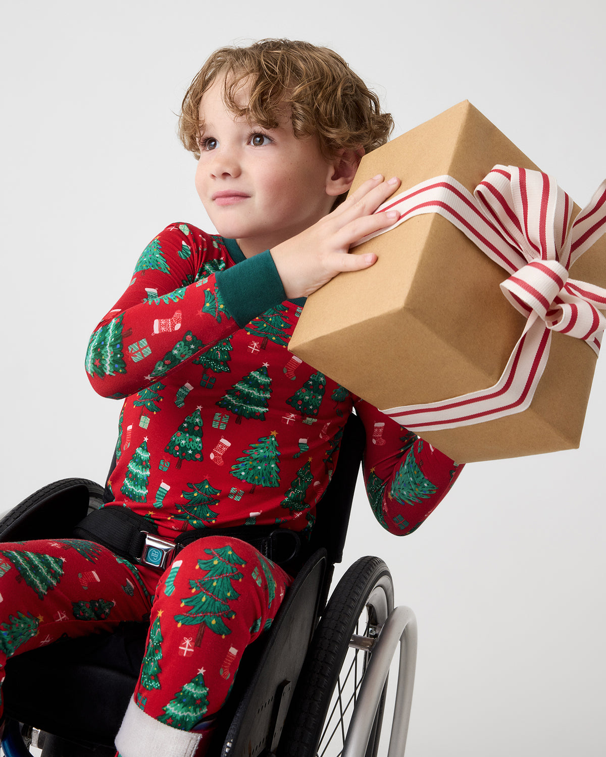A young child in a wheelchair wearing red Christmas pajamas with a green tree pattern, holding a large brown gift box tied with a red and white striped ribbon against a light gray background.