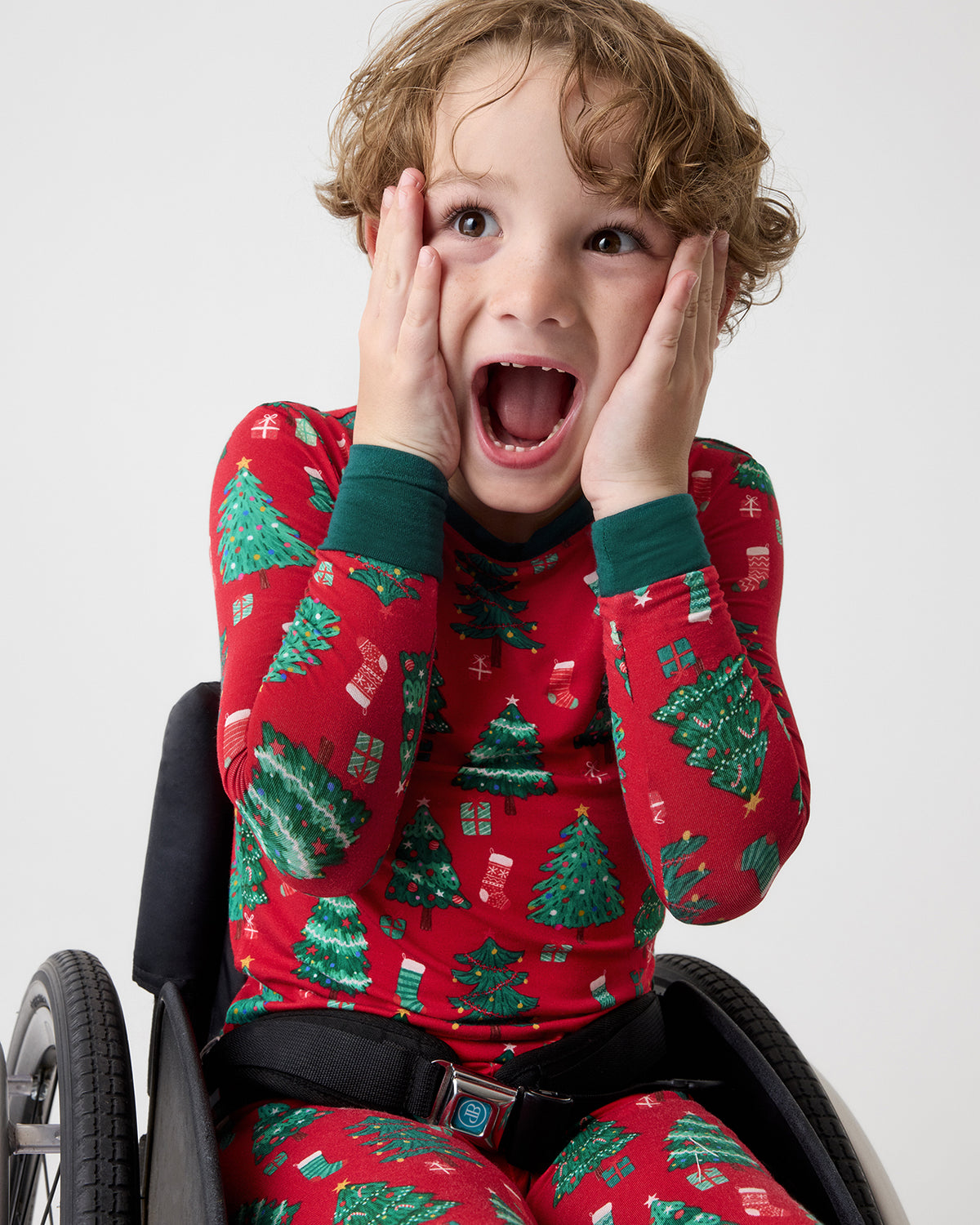 A young child sits in a wheelchair, wearing a red long-sleeve pajama set with a green Christmas tree and gift pattern. 