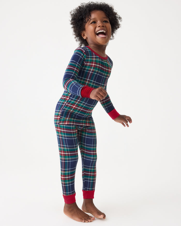 A young child wearing a long-sleeve plaid pajama set with red cuffs and collar, standing barefoot against a plain white studio background.