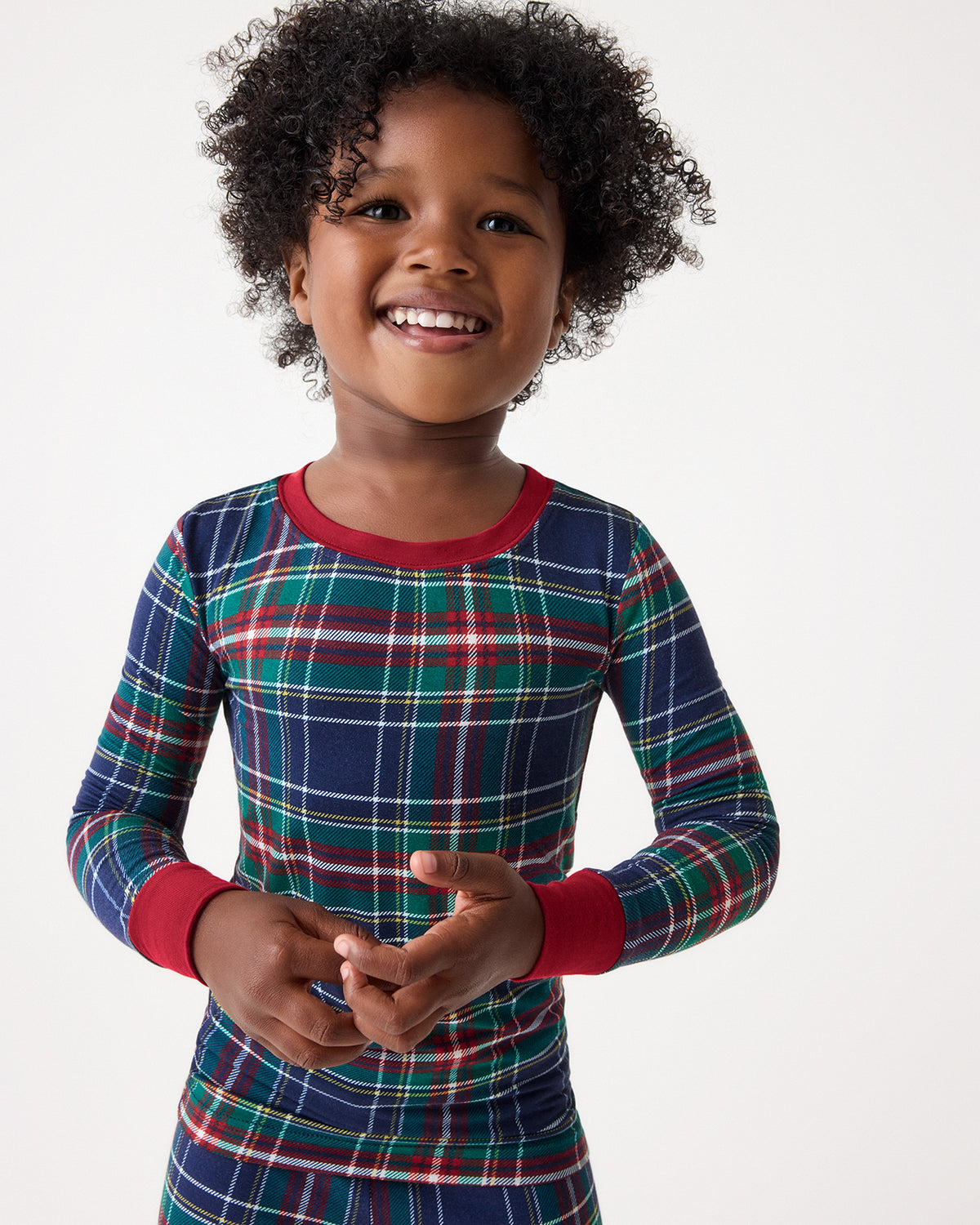 A smiling young child wearing a long-sleeve plaid pajama top in navy, green, red, and white, with solid red collar and cuffs, photographed against a plain light background.