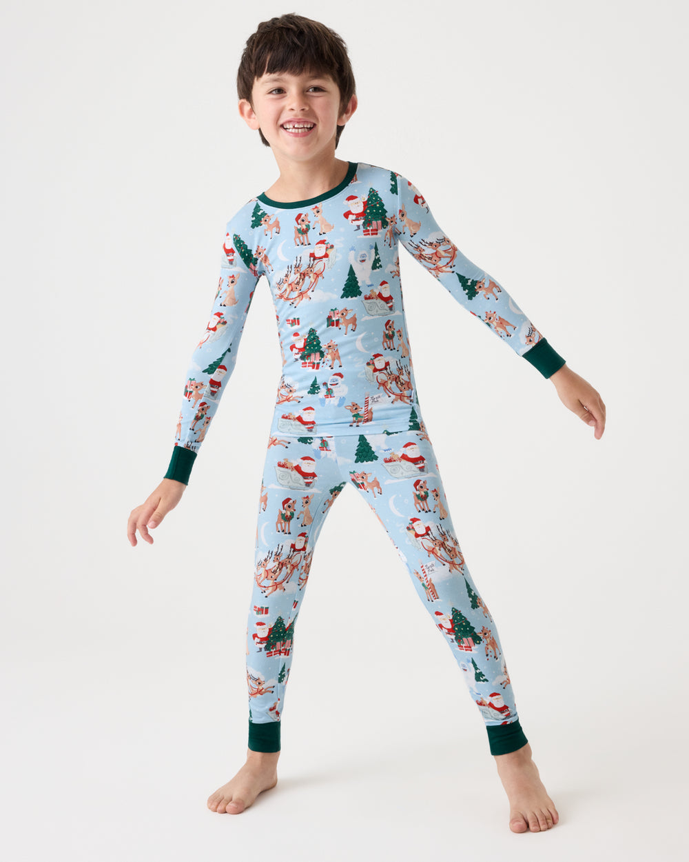 A young boy smiling and posing in a light blue long-sleeve pajama set with a festive Christmas print featuring Santa Claus, snowmen, and trees, set against a plain white studio background.