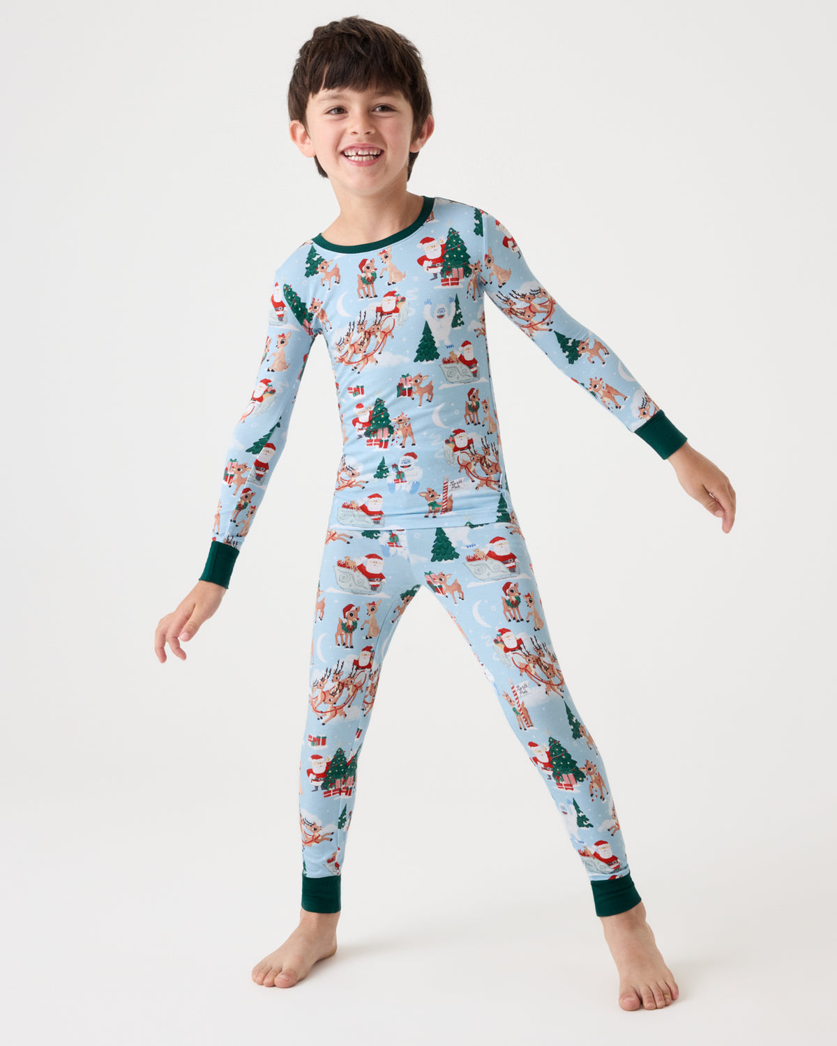 A young boy smiling and posing in a light blue long-sleeve pajama set with a festive Christmas print featuring Santa Claus, snowmen, and trees, set against a plain white studio background.