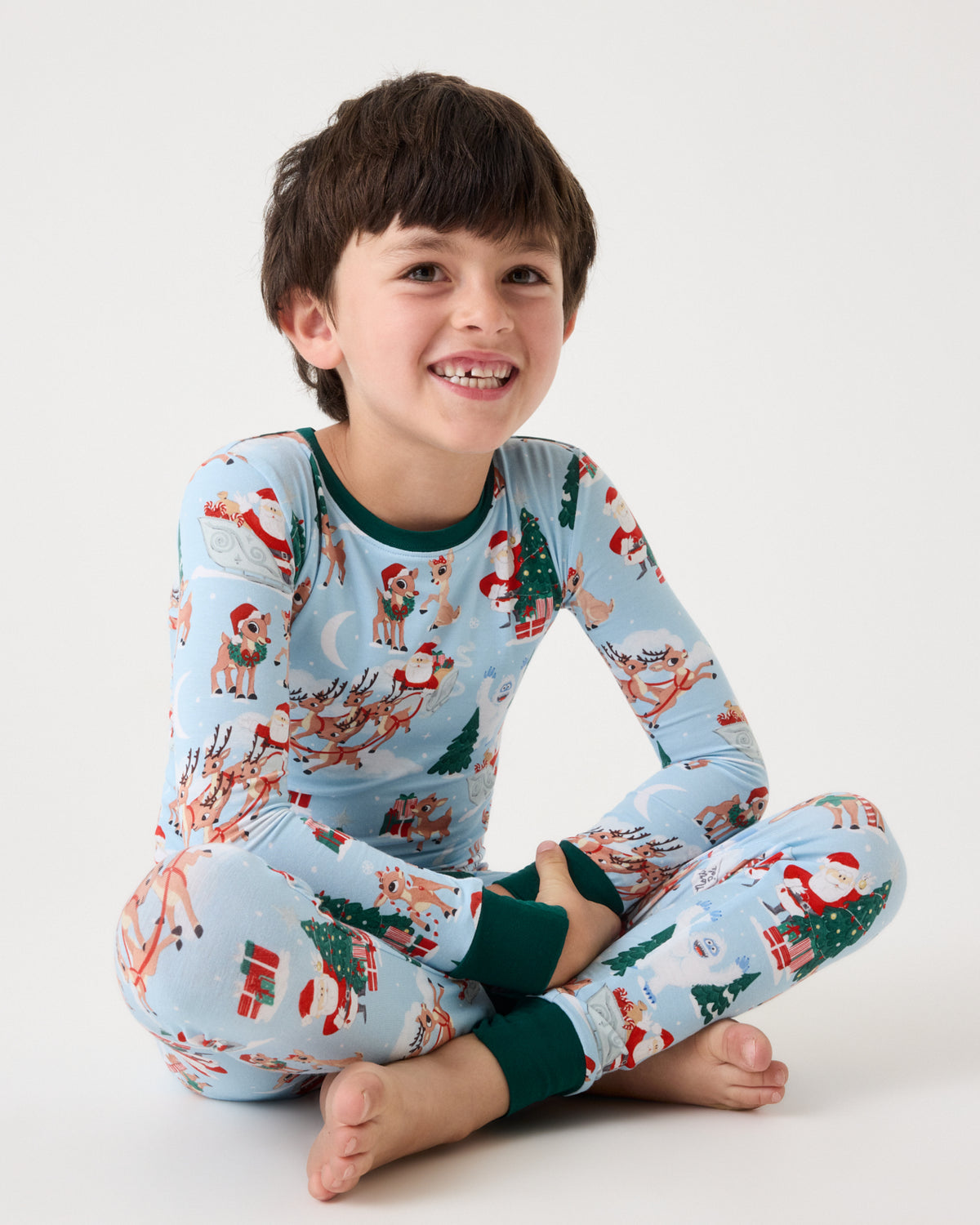 A young boy smiling while sitting cross-legged, wearing a light blue long-sleeve pajama set with a festive Christmas print of Santa Claus, reindeer, and trees, with dark green cuffs and collar.