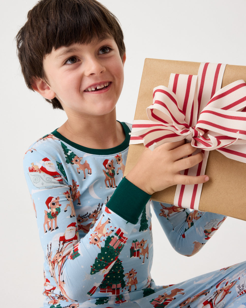 A smiling young boy wearing light blue Christmas pajamas with a green collar and cuffs, holding a brown gift box tied with a red and white striped ribbon against a plain white background.