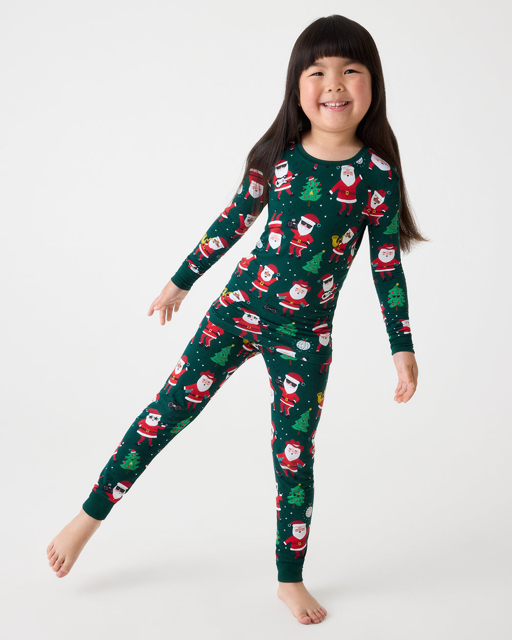 A young girl smiling and posing in a dark green Christmas-themed long-sleeve pajama set with an all-over Santa Claus and Christmas tree pattern, set against a plain light gray studio background.