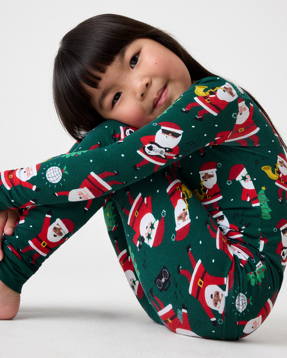 A young girl with dark hair sits curled up in a circle, wearing a dark green long-sleeve pajama set with a festive Santa Claus print. The background is a clean, light gray studio backdrop.