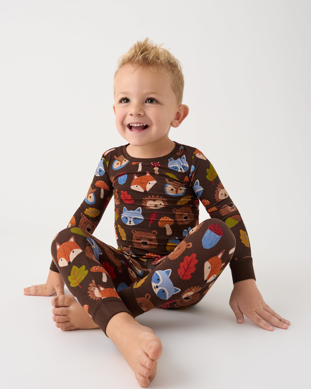 A smiling toddler sitting cross-legged on a light gray studio background, wearing a long-sleeve matching pajama set with a brown base and a colorful woodland print of foxes, mushrooms, acorns, and leaves.