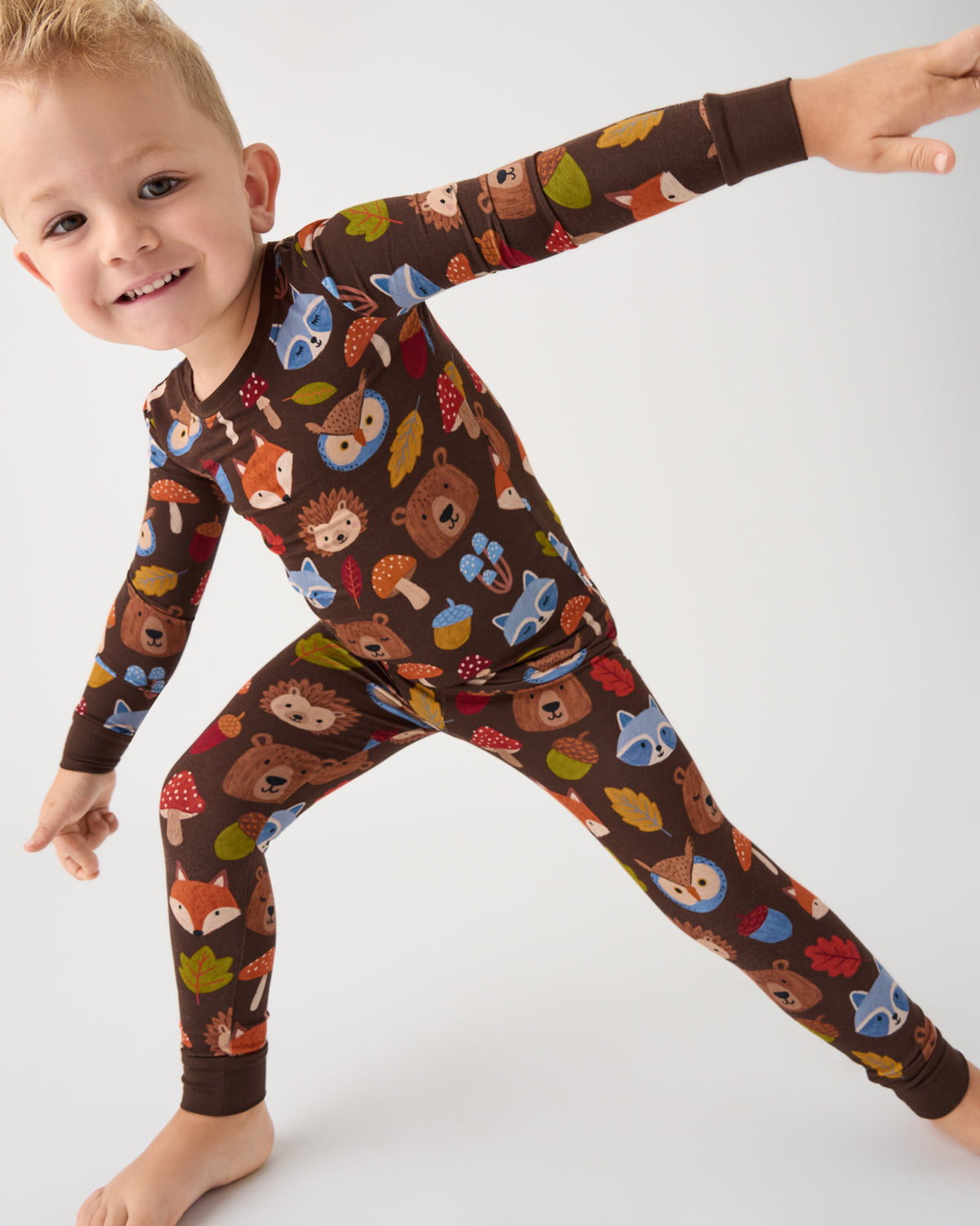 A smiling young child wearing a long-sleeve one-piece pajama set with a woodland forest print, posed dynamically against a clean white studio background.