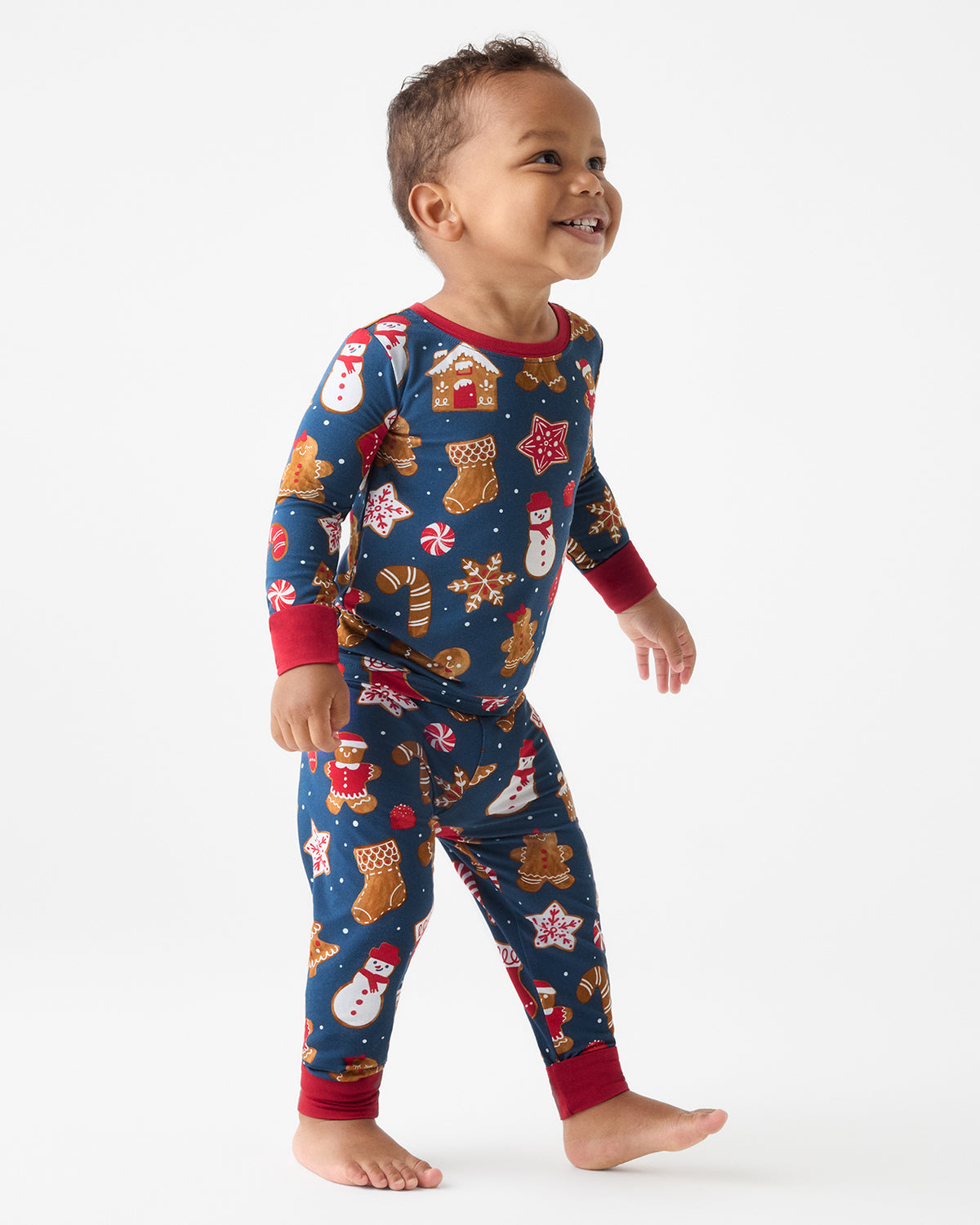 A toddler walking barefoot on a light gray studio background, wearing a long-sleeve Christmas-themed pajama set with a navy blue base and red cuffs and collar.