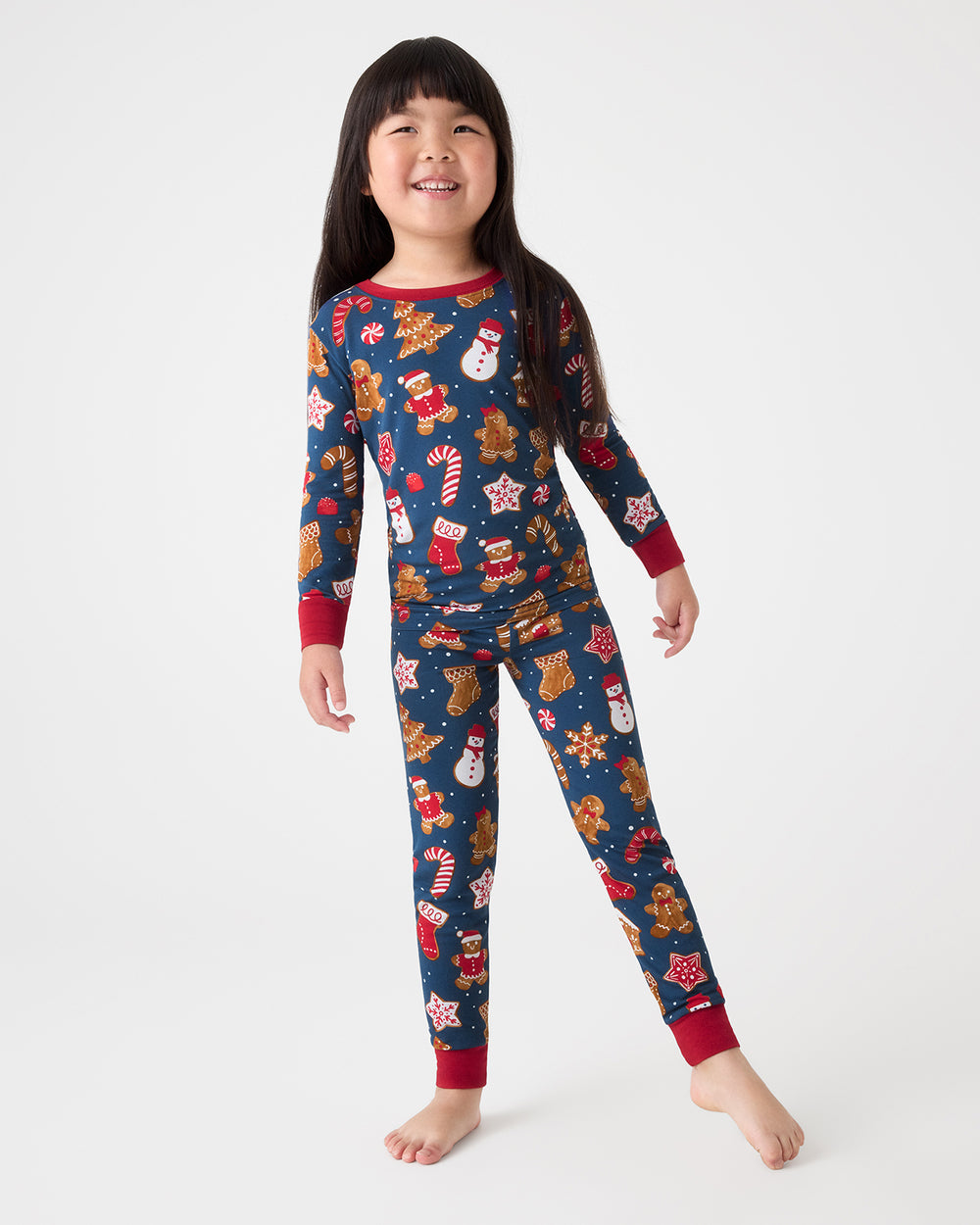A young girl modeling a long-sleeve Christmas-themed pajama set with a navy blue background and festive holiday prints, set against a plain light gray studio background.