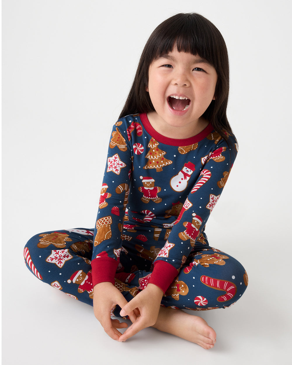 A young girl sits cross-legged on a light gray studio background, wearing a navy blue Christmas-themed long-sleeve pajama set with red cuffs and a festive print of gingerbread cookies, Santa Claus, snowmen, and candy canes.