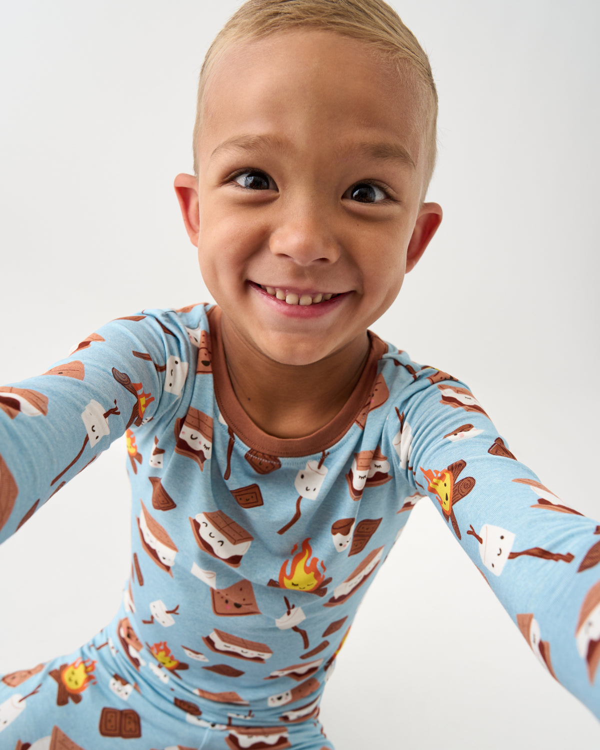 A smiling young child wearing a light blue long-sleeve pajama top with a s'mores print, photographed against a plain light gray background.
