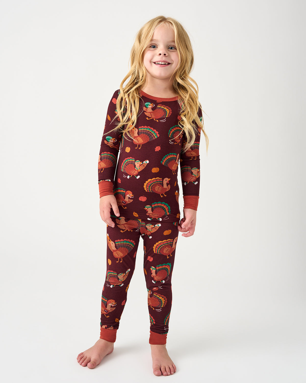 A young girl with long blonde hair stands barefoot against a light gray studio background, wearing a long-sleeve pajama set with a dark red base and an all-over autumn turkey and feather print.